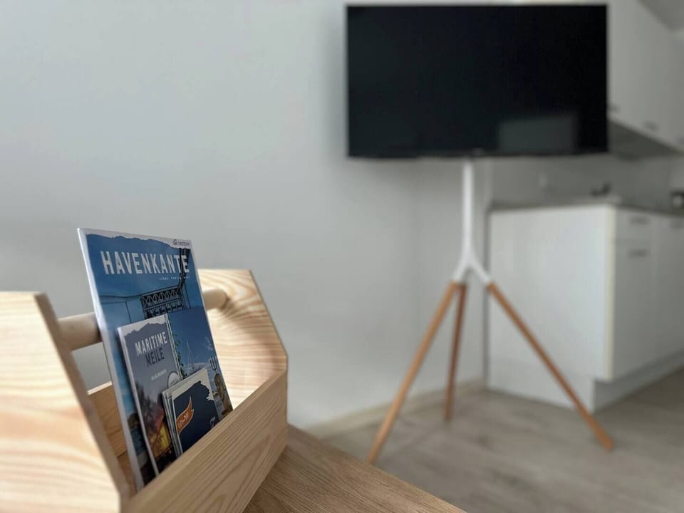 Detail mit Willkommenskorb und lokalen Broschüren; im Hintergrund moderner TV auf Stativ in der offenen Wohnküche – praktische Infos treffen auf stylische Ausstattung.