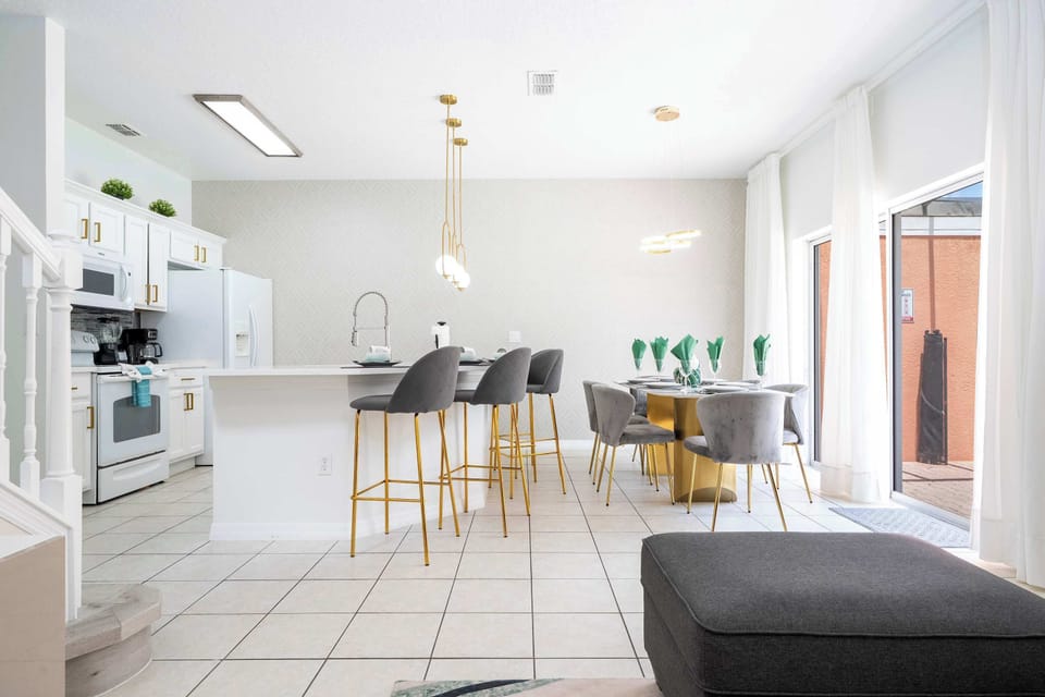 Whip up a feast in this bright, all-white kitchen with a sleek island and four plush gray bar stools. Gold-accent pendant lights & a chic dining table complete the space-perfect for morning coffee or family meals!