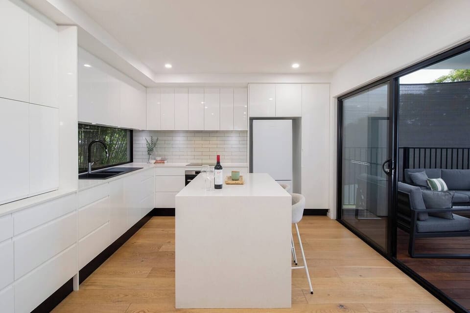 Prepare family meals or share a bottle of wine at the stone island bench in the full-sized kitchen, equipped with modern appliances, an induction cooktop, and a large window framing the bamboo garden.