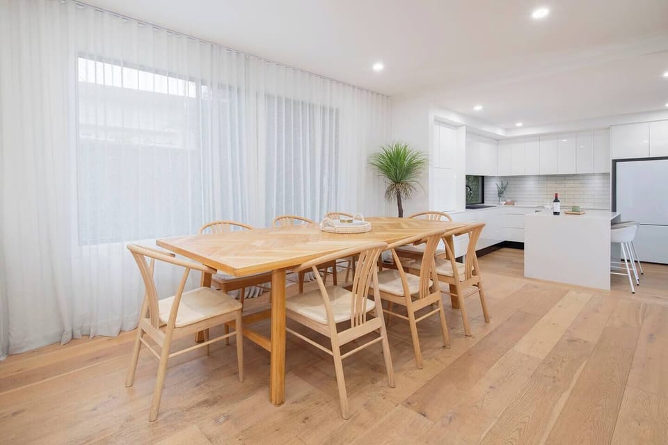 Dine together in the inviting dining area, where woven chairs and a timber table reflect the home’s coastal palette, just steps from the kitchen and main entry.