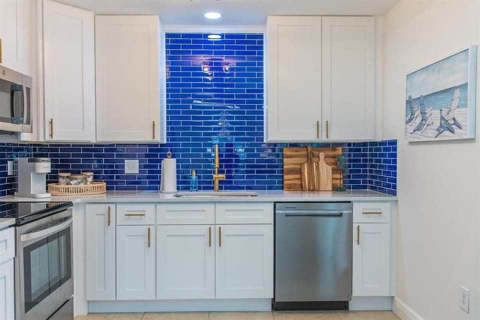 Fully updated kitchen with stainless steel appliances, white cabinetry, and bold blue tile backsplash. Includes a large prep island, open shelving, and bright window views.