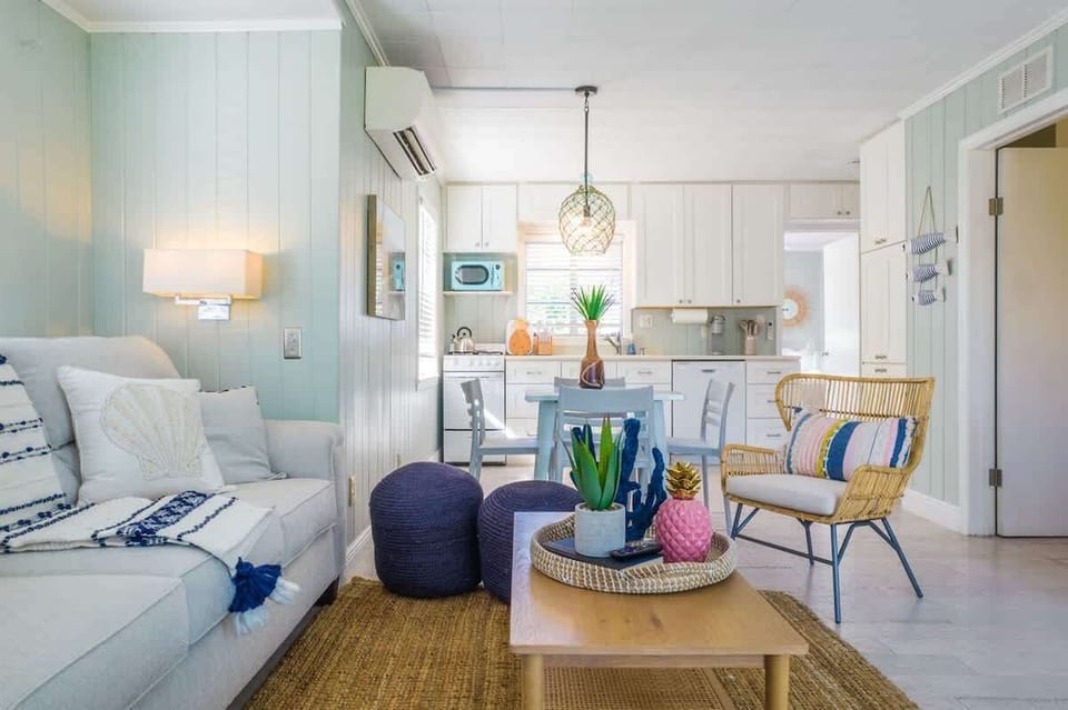 Living space features a plush sofa, accent chairs, TV, and a coffee table. The open layout connects to a kitchen with full appliances, dining table, and stylish pendant lighting.