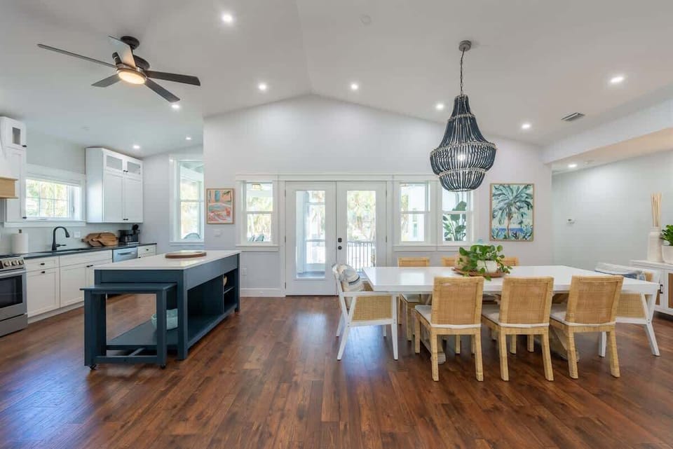 Bright dining area with a long table, rattan chairs, and a statement black chandelier. Open layout leads to a modern kitchen with stainless steel appliances, white cabinetry, and island seating, all framed by wood floors and high ceilings.