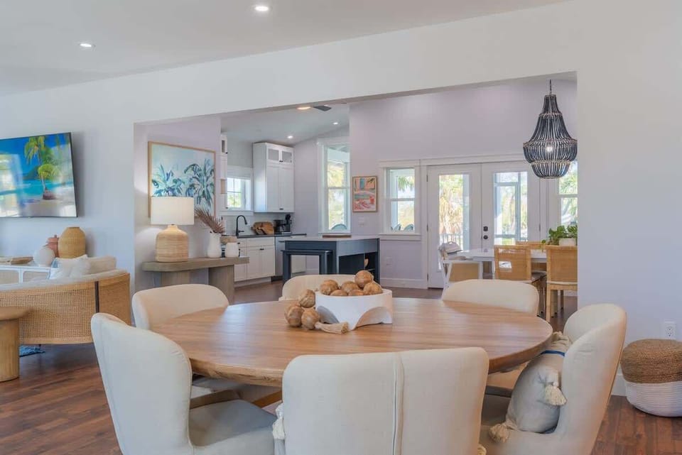 Dining area with a round wood table, cushioned chairs, and soft ambient lighting. Flows into a bright open-concept layout with hardwood floors, ceiling fans, modern fixtures, and access to kitchen, living, and outdoor spaces.
