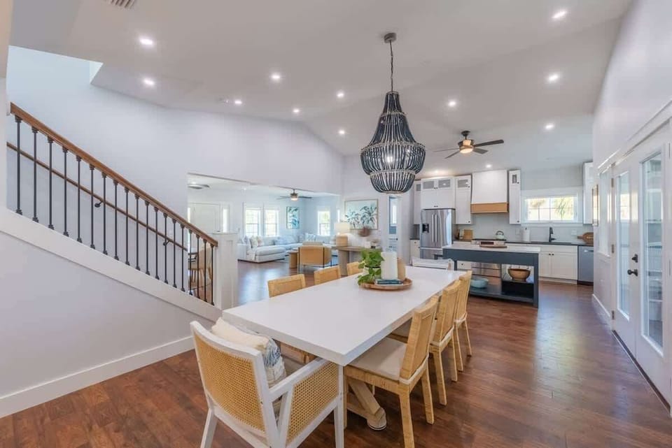 Bright dining area with a long table, rattan chairs, and a statement black chandelier. Open layout leads to a modern kitchen with stainless steel appliances, white cabinetry, and island seating, all framed by wood floors and high ceilings.