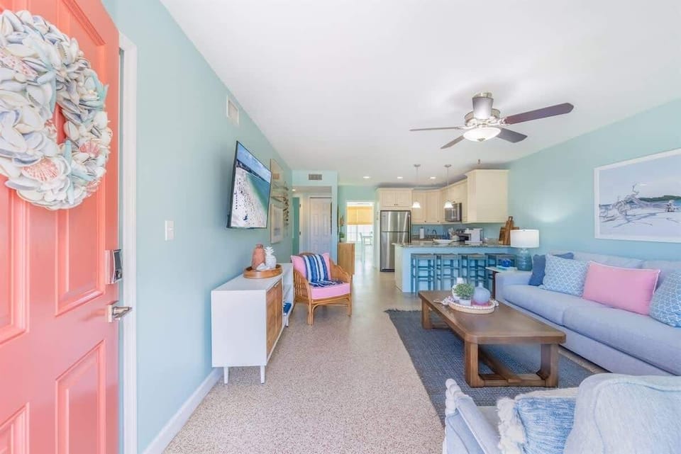 Open-concept living room with a plush sofa, accent chairs, a smart TV, and a wood coffee table. Coastal decor, pastel tones, and woven accents complete the relaxed vibe.