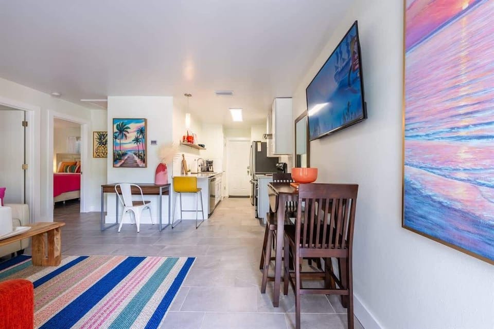 Living area with a leather sofa, bright accent chairs, and a wall-mounted TV. Includes a dining nook, workspace, and open kitchen.