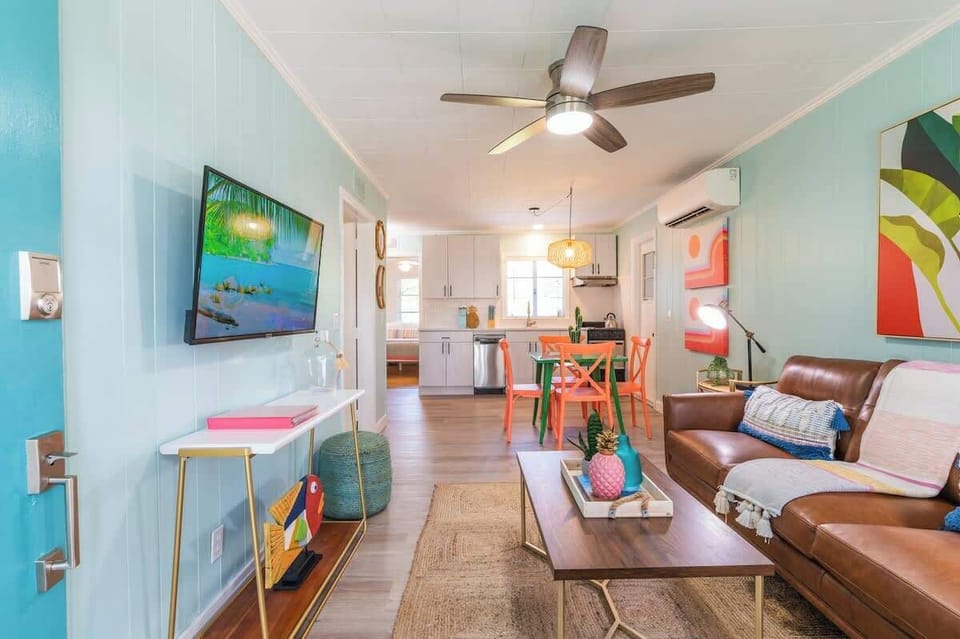 Tropical living room with leather seating, rattan accents, a smart TV, ceiling fan, bright decor, and open layout to the kitchen and dining area.