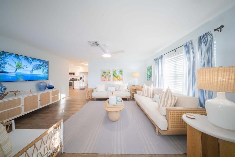 Spacious living area with sofas, rattan chairs, a large smart TV, modern coastal décor, and ceiling fan.