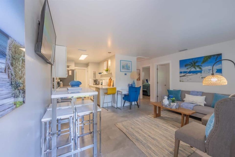 Open-plan living space with a full kitchen, breakfast bar, and dedicated workspace. Features modern appliances, comfy seating, and beachy decor throughout. Includes a mounted TV, dining nook, and vibrant accent furniture.