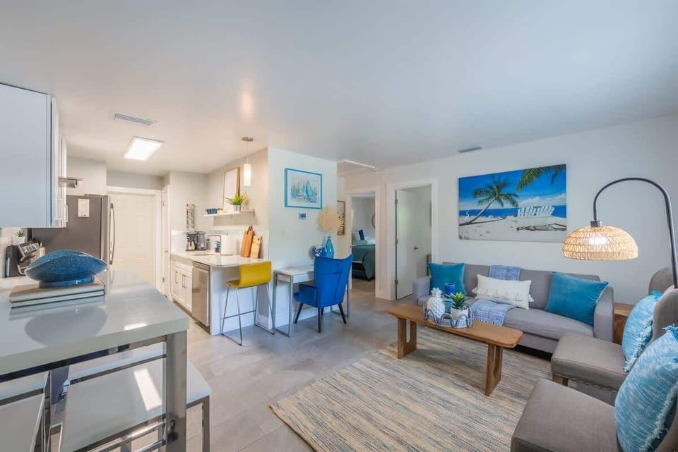 Open-plan living space with a full kitchen, breakfast bar, and dedicated workspace. Features modern appliances, comfy seating, and beachy decor throughout. Includes a mounted TV, dining nook, and vibrant accent furniture.