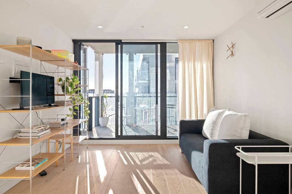 Unwind in the sunlit living area, where a deep navy sofa faces a TV framed by books and greenery, with sweeping city views beyond the glass.