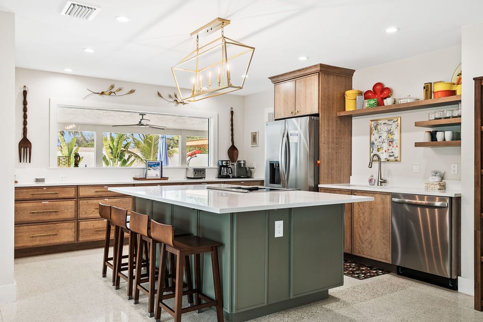 Your dream kitchen awaits with warm wood cabinets, gleaming quartz counters, and a stunning green island perfect for morning coffee or evening cocktails. Natural light pours through windows revealing tropical palms beyond.