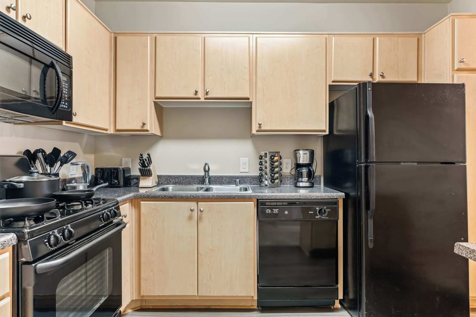 Modern kitchen with black appliances, including an oven, microwave, dishwasher, and coffee maker. Natural wooden cabinets and a granite countertop complete the contemporary style.