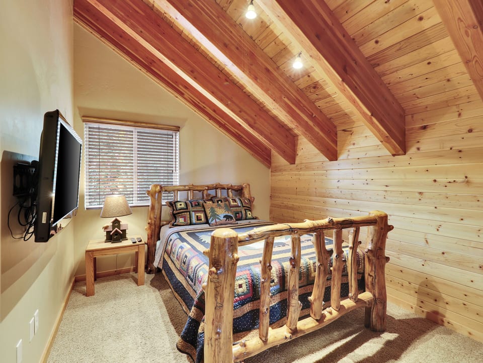 Queen bedroom with rustic log bed frame, vaulted wood-beam ceiling, flat-screen TV, and cozy mountain décor at Aspen Lodge, Brian Head, Utah. One of five bedrooms in this luxury large-group cabin sleeping 25.