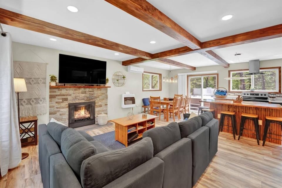 Cozy open-concept living area featuring a fireplace, wall-mounted TV, and comfortable seating. The space flows into a fully equipped kitchen with counter seating and a dining table for group meals. 