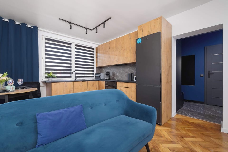 A living room and kitchen combo featuring a turquoise sofa, black-and-wood kitchen, and large windows with blinds. The open layout makes the room feel spacious.