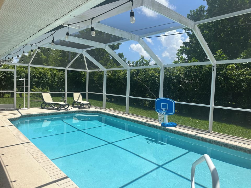 Heated pool within the screened enclosure with lounge chairs on the deck and lush greenery surrounding