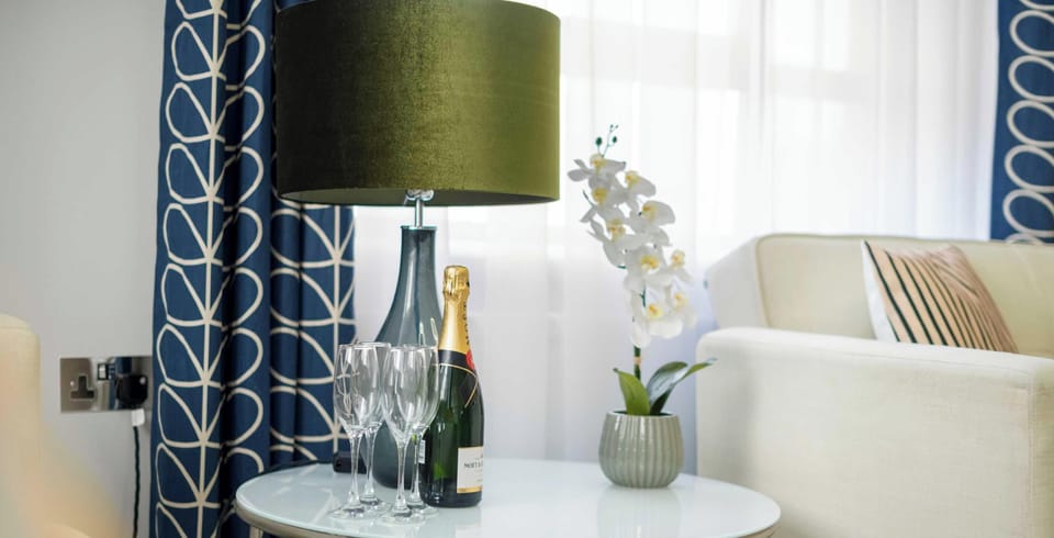 A sophisticated side table is set with a bottle of champagne and elegant glassware, framed by a deep green lamp and patterned blue curtains.