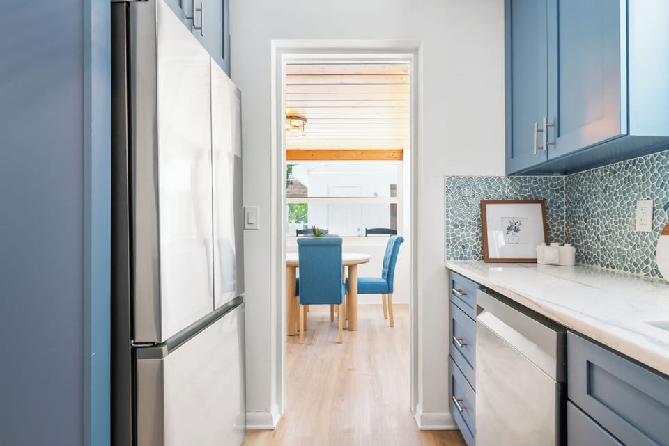Kitchen passthrough to sunlit dining nook — stainless fridge, mosaic backsplash, and a direct sightline to the covered dining area beyond