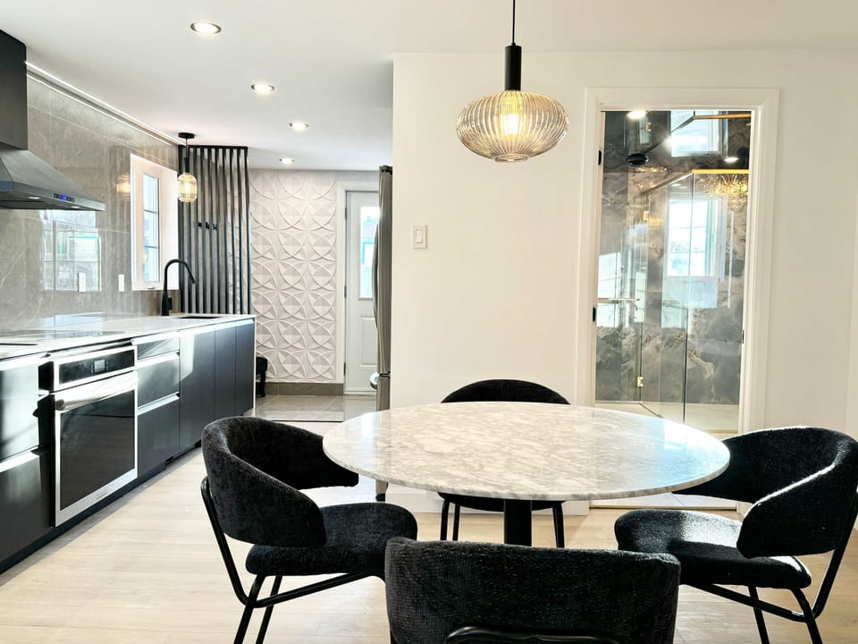 Un espace moderne et épuré à aire ouverte, doté d'un coin repas avec une table ronde à plateau de marbre et quatre chaises sombres. Une cuisine contemporaine avec des armoires foncées laquées est visible à l'arrière-plan.