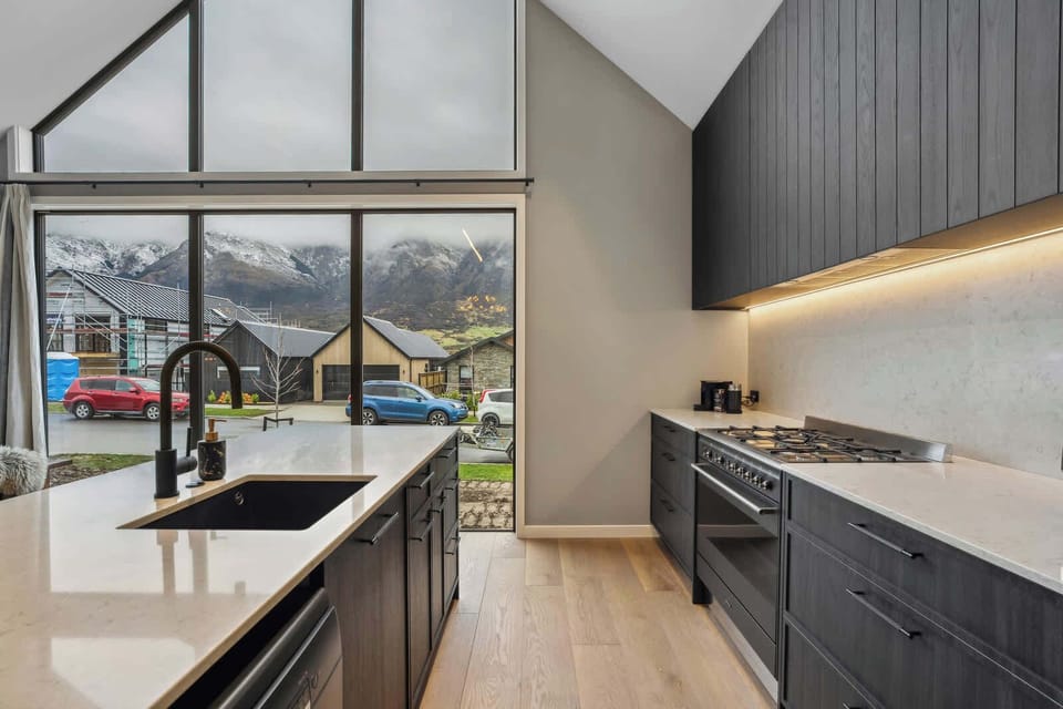 Spacious open-plan kitchen and dining area with soaring ceilings, warm timber floors, and mountain views through oversized windows. Ideal for shared meals and family moments.