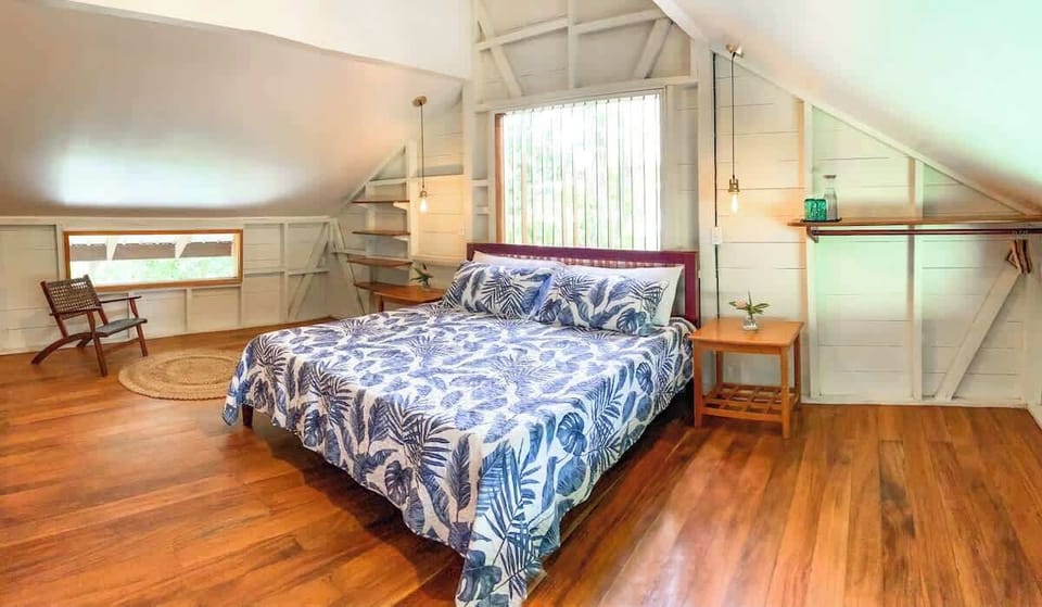 Relax in this cozy loft-style bedroom with tropical charm. Enjoy natural light, warm wooden floors, and a plush king bed with soft linens. A peaceful jungle retreat designed for comfort and tranquility.
