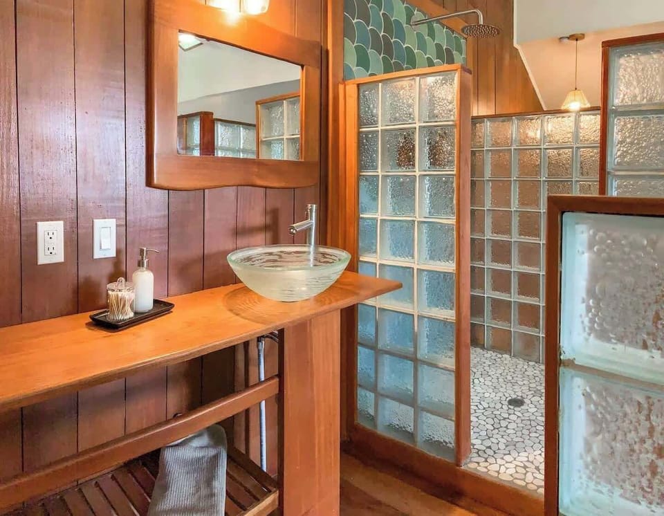 Enjoy a spa-like experience in this rustic yet modern bathroom. Featuring a stylish glass vessel sink, heated rain shower, and elegant glass block walls, this space blends natural wood warmth with refreshing luxury.