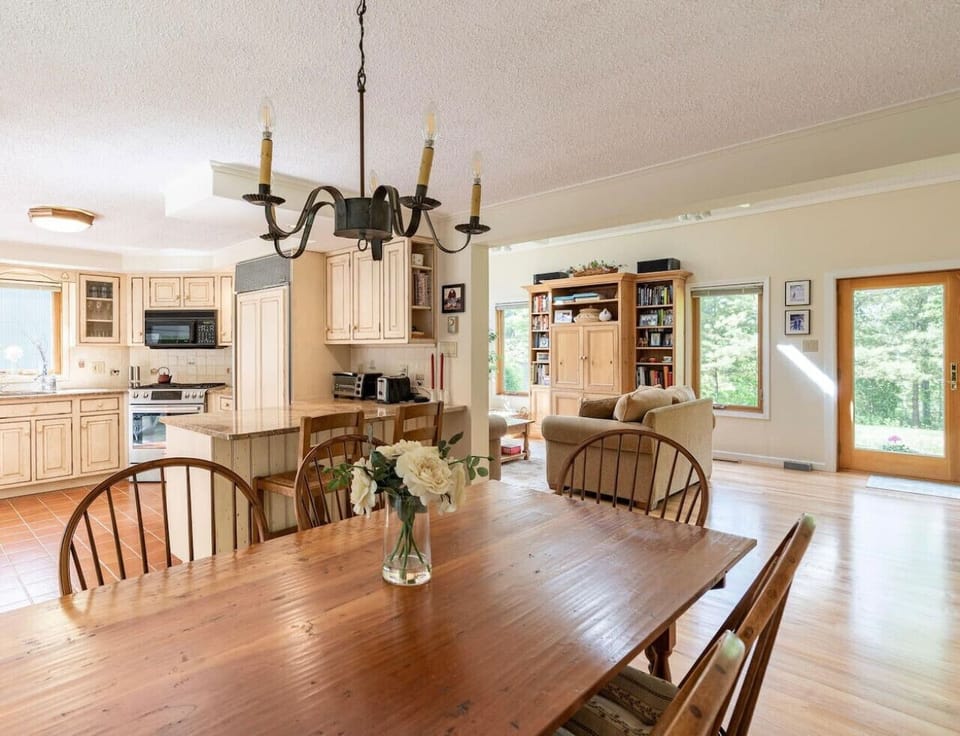 Dining table with wooden chairs for 6, chandelier lighting, and view into kitchen and living room.