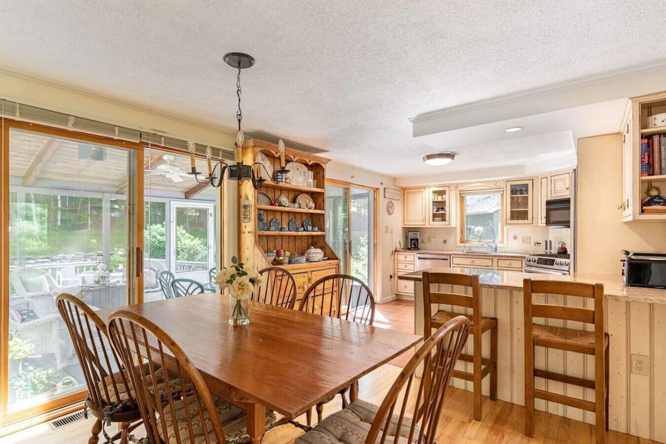 Dining area with large windows overlooking greenery and adjacent to kitchen.
