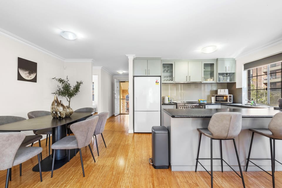 Dine in a modern space featuring a gray dining table and chairs, seamlessly connected to a bright kitchen with gray cabinetry and a refrigerator, offering a comfortable area.