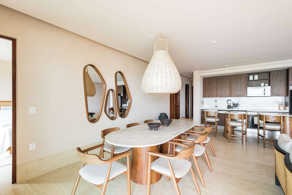 A bright dining area with an oval table, wooden chairs, and a woven pendant light, adjacent to a modern kitchen.
