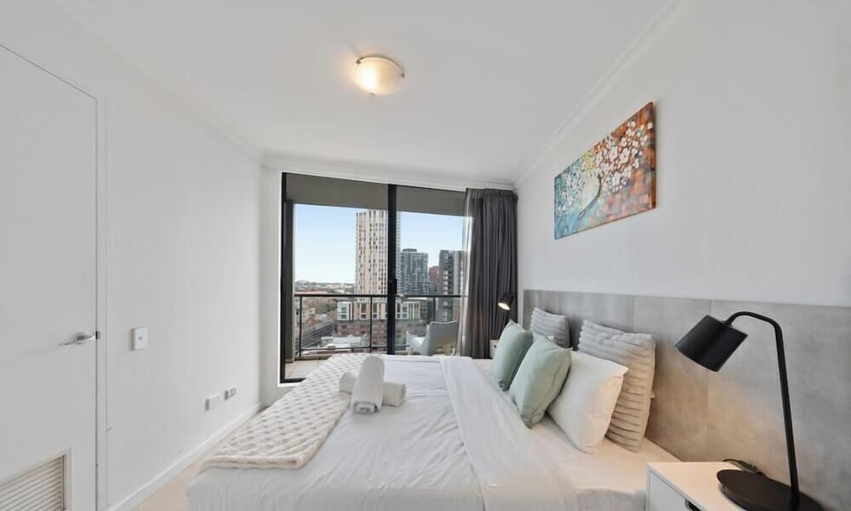 A spacious, bright bedroom featuring a large bed with crisp white linens, light green accent pillows, and a contemporary grey headboard.