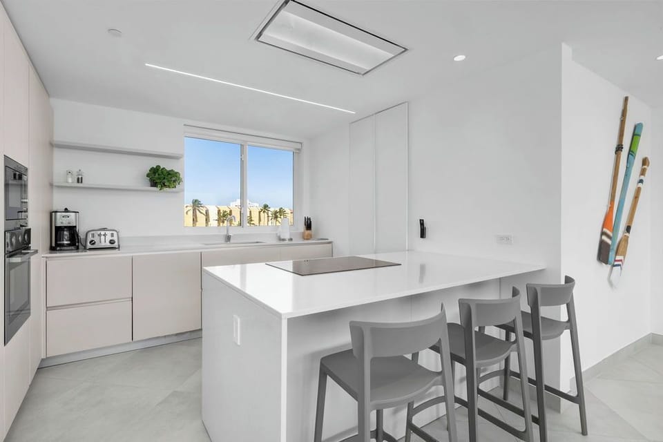 Your sleek modern kitchen features stainless appliances, a spacious island with bar seating, and bright natural light streaming in through large windows overlooking palm-lined surroundings.