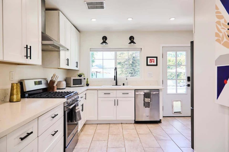This modern kitchen features white shaker cabinets, black hardware, and stainless steel appliances, including a gas range and dishwasher. A large window brightens the space, and the back door offers convenient access to the outdoors.