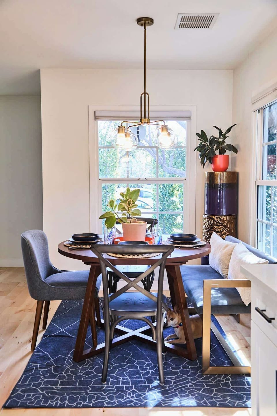 A bright dining nook with a round wooden table, upholstered chairs, and a built-in bench accented by patterned pillows. Large windows bring in natural light, while potted plants and a modern pendant light create a warm, inviting atmosphere.
