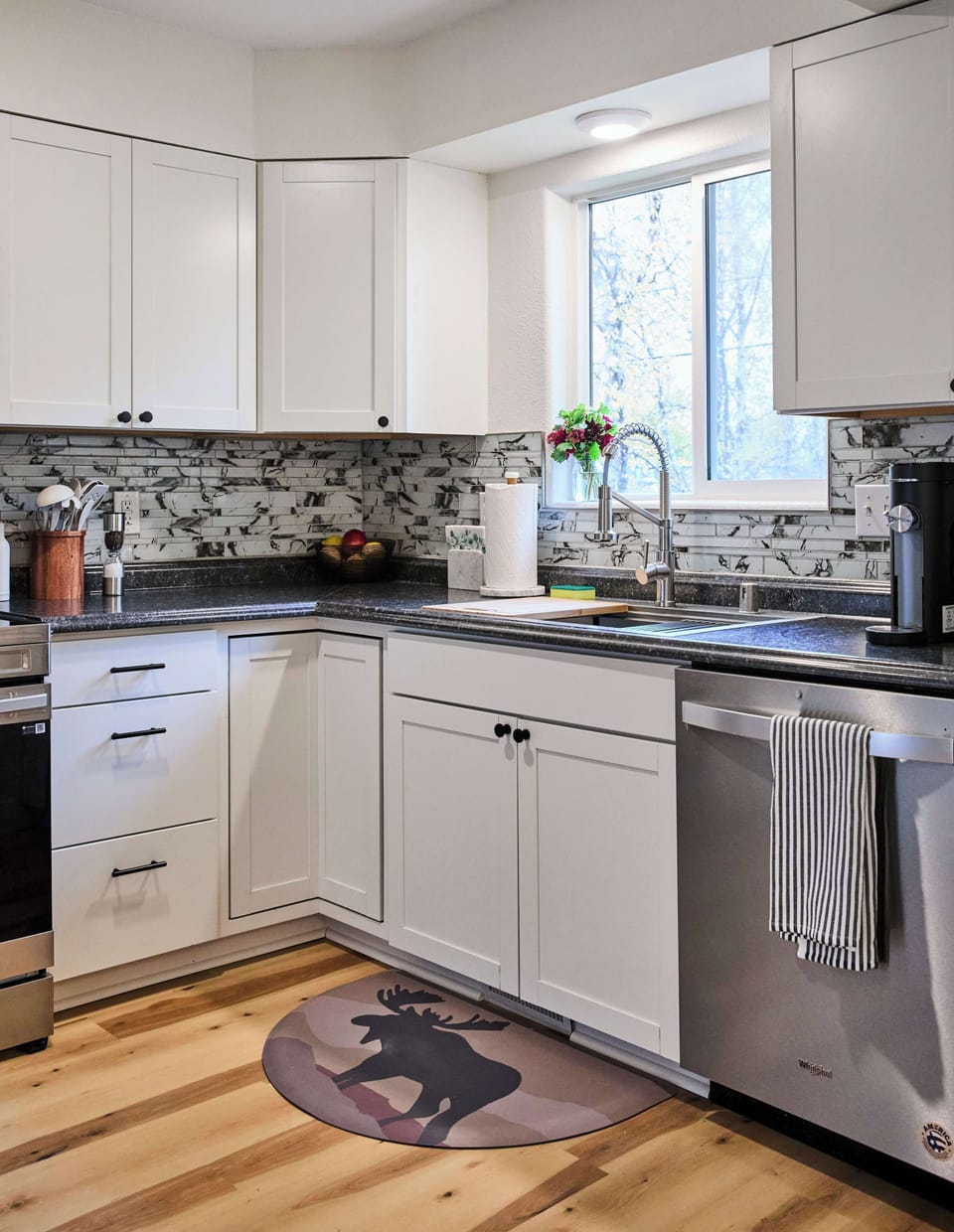 This kitchen features sleek white cabinetry, stainless steel appliances, and a modern mosaic tile backsplash. Natural light brightens the space, highlighting warm wood floors and a playful bear rug for a cozy, welcoming touch.