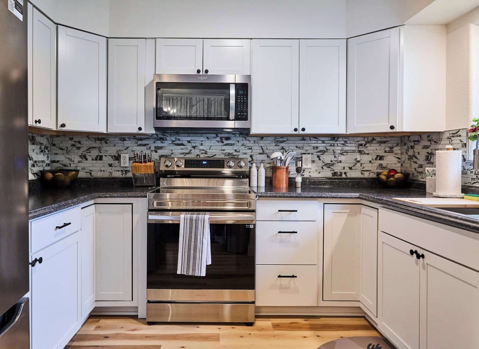 This modern kitchen showcases stainless steel appliances, white cabinetry, and a striking mosaic tile backsplash. The wood flooring adds warmth, while sleek finishes create a functional and stylish cooking space.