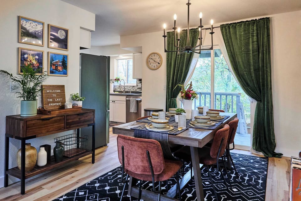 Bright dining space with a rustic wooden table, warm leather chairs, and a bold chandelier. Green curtains and sliding doors bring in natural light, while a sideboard with decor and framed art adds charm for gatherings.