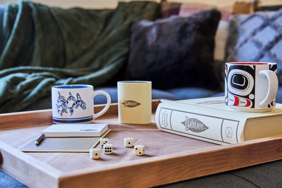 A cozy coffee table tray setup with mugs, a notebook, dice, and a book creates the perfect scene for a relaxed morning or game night, set against a backdrop of soft blankets and pillows.