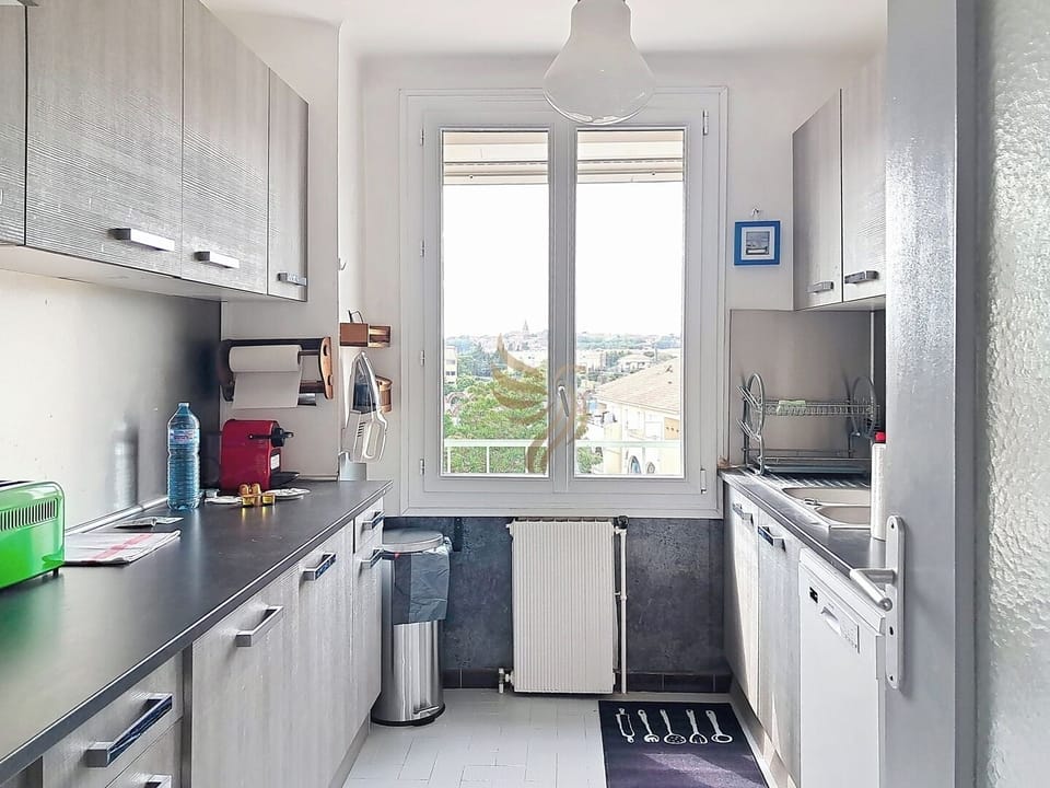 Une cuisine lumineuse, compacte et fonctionnelle avec des placards modernes en bois gris et des équipements de base. Une grande fenêtre illumine l'espace en montrant une vue dégagée.