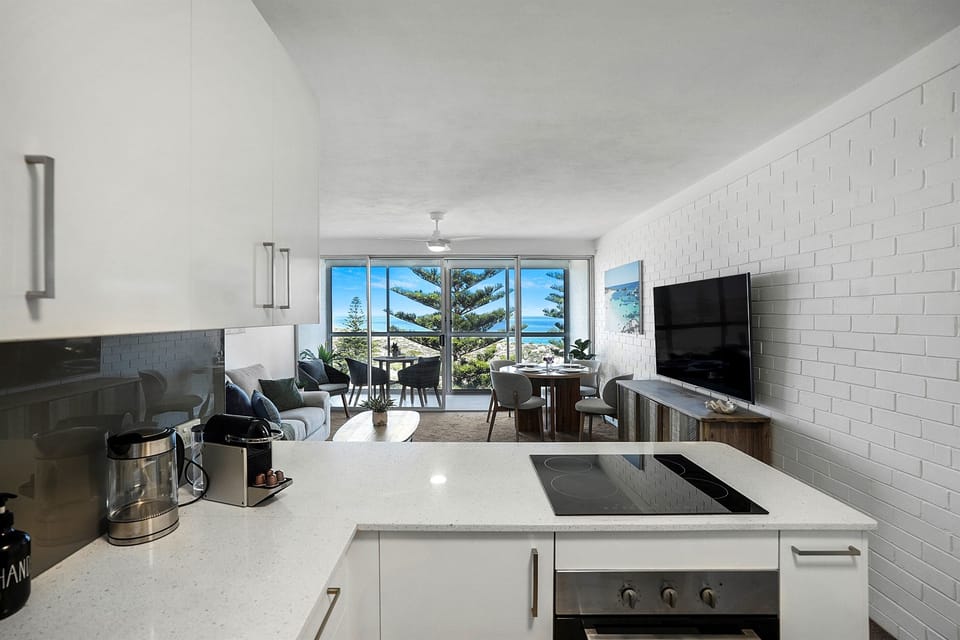 Seamless flow from kitchen to living area with a flat-screen TV and panoramic windows overlooking the coastline.