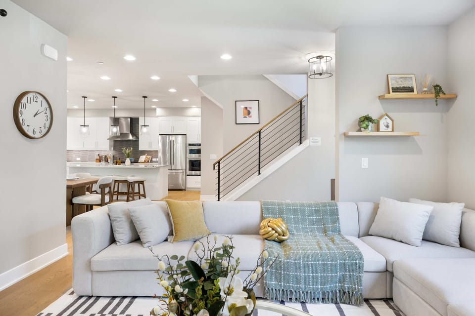 A neutral L-shaped sofa with teal-and-gold accents faces a minimalist kitchen with pendant lighting. The black-and-wood staircase adds contrast.