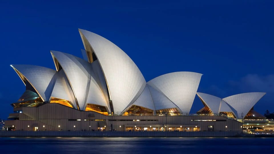 The iconic white sails of the Sydney Opera House glow brilliantly against a deep blue twilight sky, casting warm golden light from within over the dark harbor waters