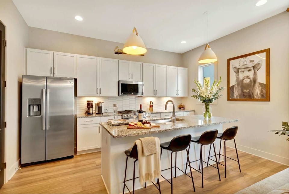 Three bar stools pull up to a granite island across from stainless steel appliances and white cabinetry, the setup for prepping snacks or cooking a full dinner for the whole group