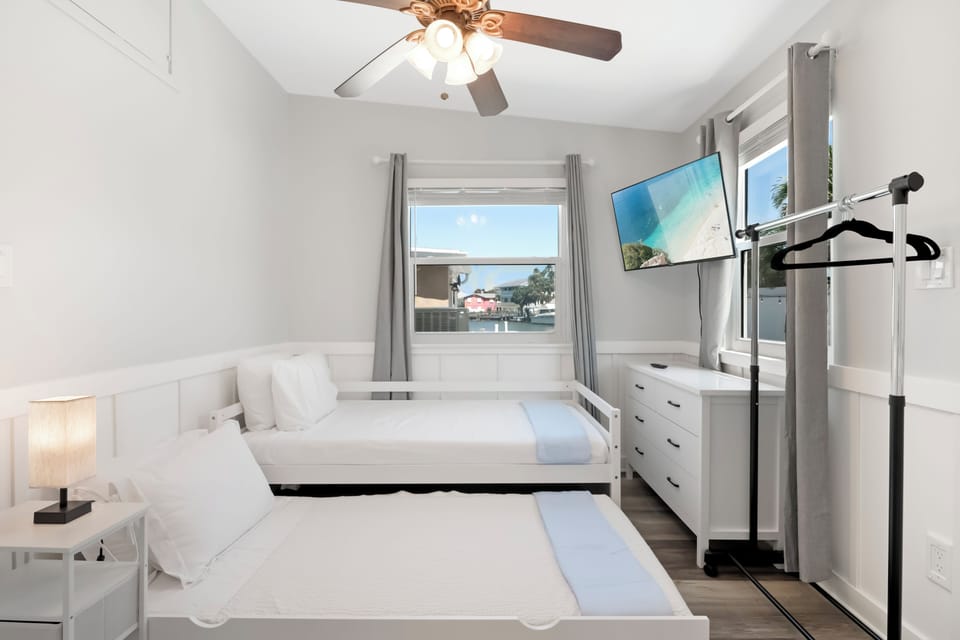 Bright guest room with two twin beds, smart TV, ceiling fan, and natural light.
