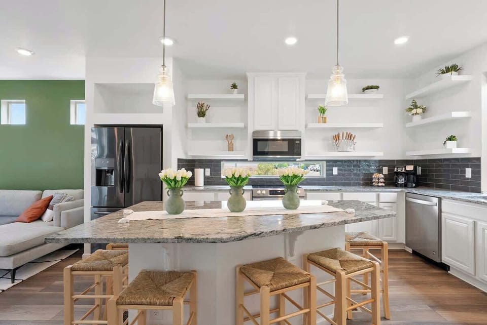 Modern kitchen with granite island, stainless steel appliances, and cozy seating for six. Open design connects to living area with green accent wall. Bright, stylish, and perfect for social gatherings. Book your stay today!
