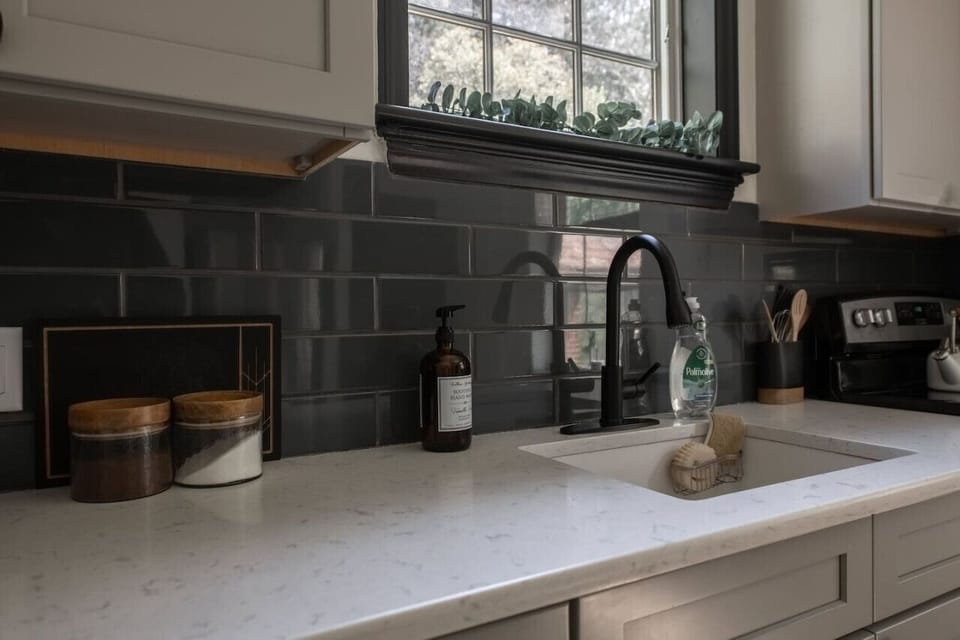 A close-up of a modern kitchen countertop, featuring a sink with a black faucet, containers, soap, and a window above