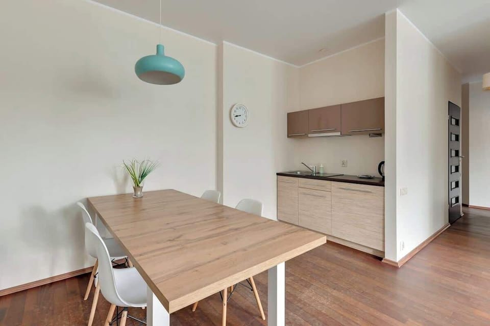 A modern kitchen area with pale wooden cabinets and a sleek dining table. A teal pendant light above the table adds a pop of color to the otherwise minimal design.