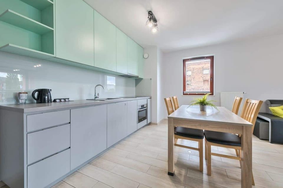 A modern kitchen with sleek light gray cabinetry and a wooden dining table set with chairs. Large windows brighten the space.